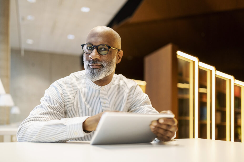 Portrait of mature man sitting at desk in a library with digital tablet